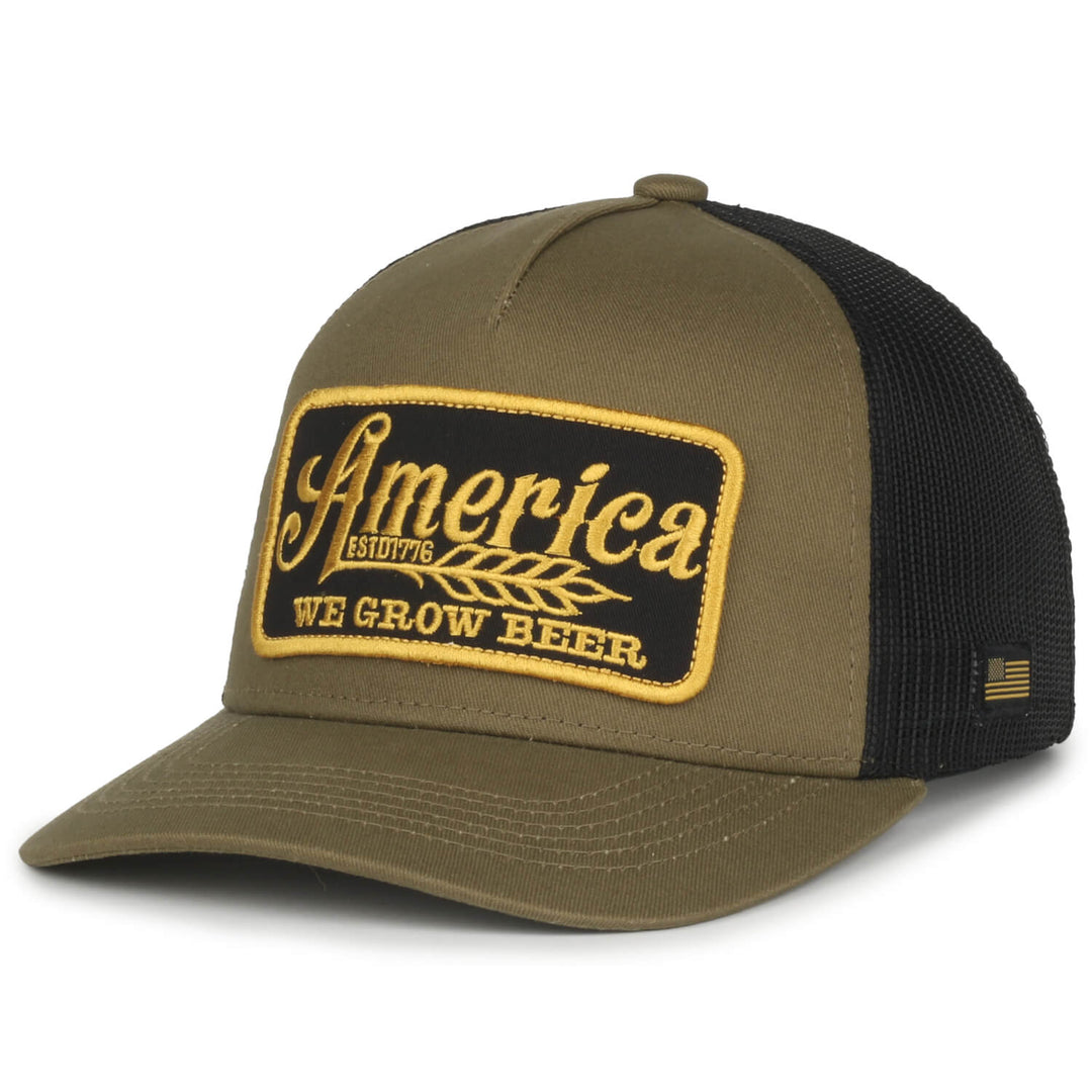 The We Grow Beer Hat-Olive by Rural Cloth features a khaki and black trucker cap design with a patch showcasing the text "America Estd 1776 We Grow Beer" in yellow and white embroidery. This hat includes a breathable mesh back, an adjustable snapback closure, and an American flag tag on the side.
