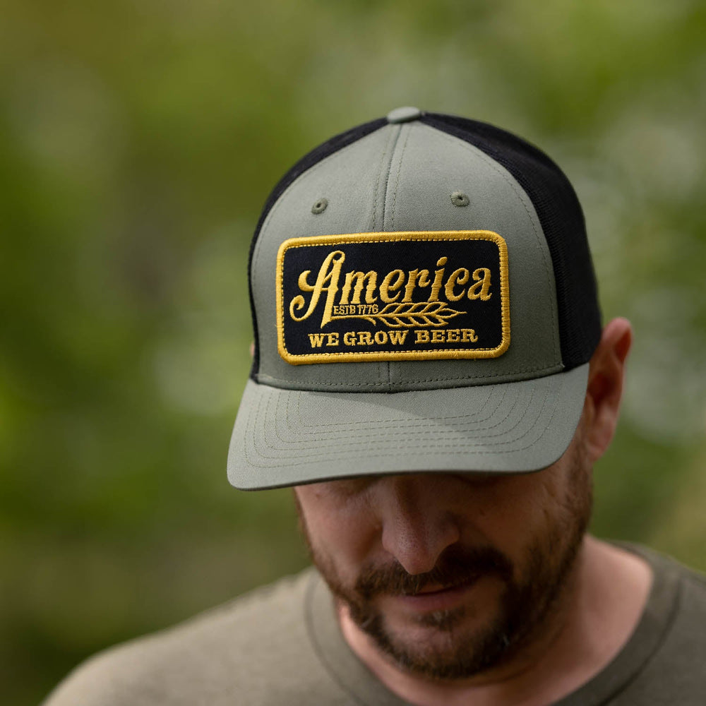 A person is wearing an olive green and black We Grow Beer Hat-Olive from Rural Cloth, featuring a yellow and black patch that reads "America: We Grow Beer" and a breathable mesh back. The person is looking down against a backdrop of blurred greenery.