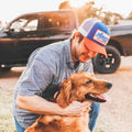 A man wearing a Rural Cloth We Grow Beer Hat in red, white, and blue kneels to pet a golden retriever. The man is outdoors in front of a black pickup truck as the sun sets in the background, epitomizing an idyllic American evening.