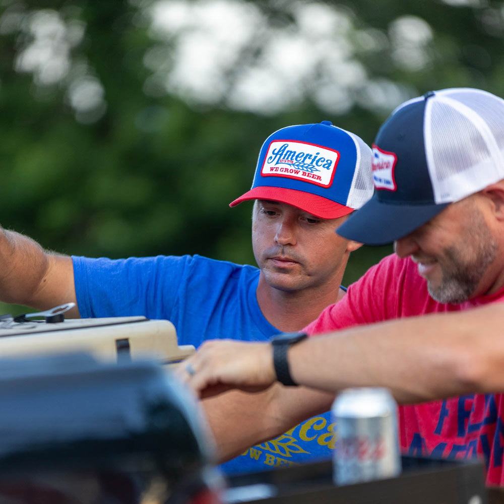 Two men are working together outdoors, epitomizing the spirit of America. They are both sporting "We Grow Beer Hat-Red, White and Blue" caps from Rural Cloth, along with t-shirts—one blue and one red. One man holds a cooler lid while the other reaches inside. A soda can is visible on the right amidst blurred greenery, capturing a slice of everyday life.