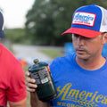 A man in a blue shirt and a We Grow Beer Hat-Red, White and Blue from Rural Cloth is holding a green beer bottle with both hands, appearing to examine it. Another person in a red shirt stands next to him, partially visible. The background is outdoors, reflecting the casual vibe of America.