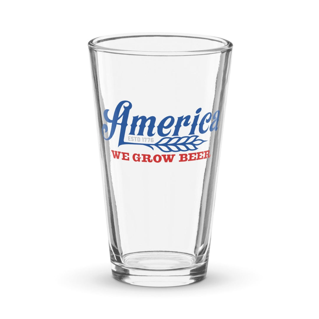 The We Grow Beer Pint Glass by Rural Cloth is a clear pint glass featuring "America" in blue script, "ESTD 1776" in small blue text beneath it, and "WE GROW BEER" in bold red uppercase letters alongside a graphic of a wheat stalk. This handmade product honors American Beer Farmers with every pint poured.