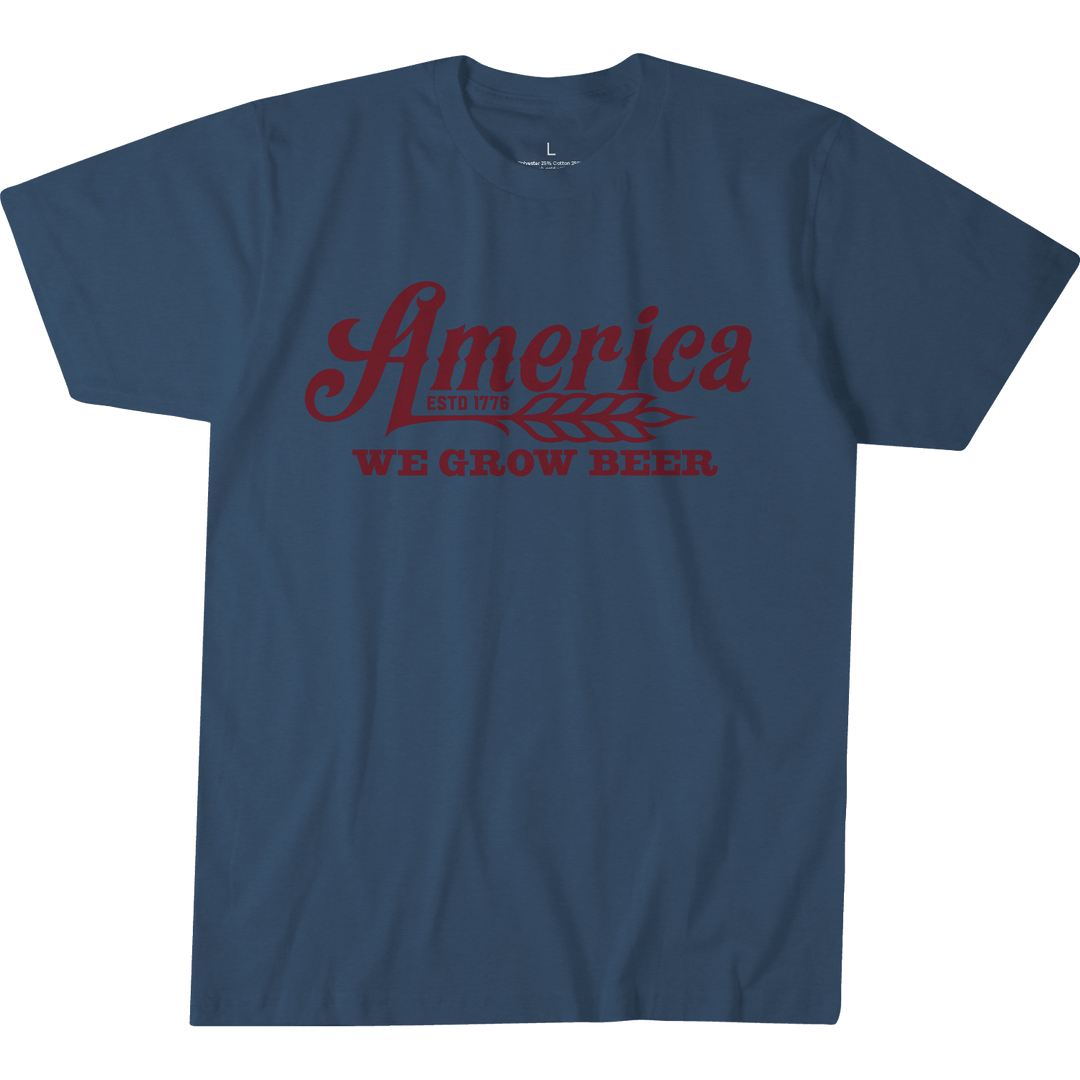 The Rural Cloth We Grow Beer Tee in Heather Blue features comfortable premium weight fabric and showcases the text "America ESTD 1776" in red, followed by an image of a wheat spike. Below, the text "WE GROW BEER" is also displayed in red. The t-shirt has a modern athletic fit and is shown flat against a plain backdrop.