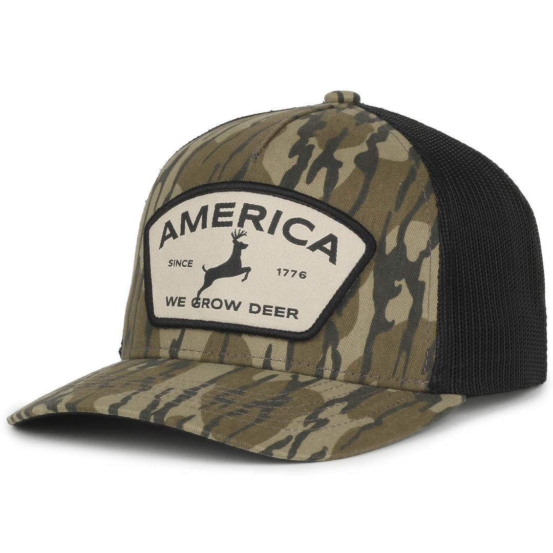 The Rural Cloth We Grow Deer Badge Hat in Bottomland features a camouflage pattern with a black mesh back and a curved brim. The patch on the front displays an image of a deer and text that reads, "AMERICA, SINCE 1776, WE GROW DEER," making it perfect for the outdoorsman.