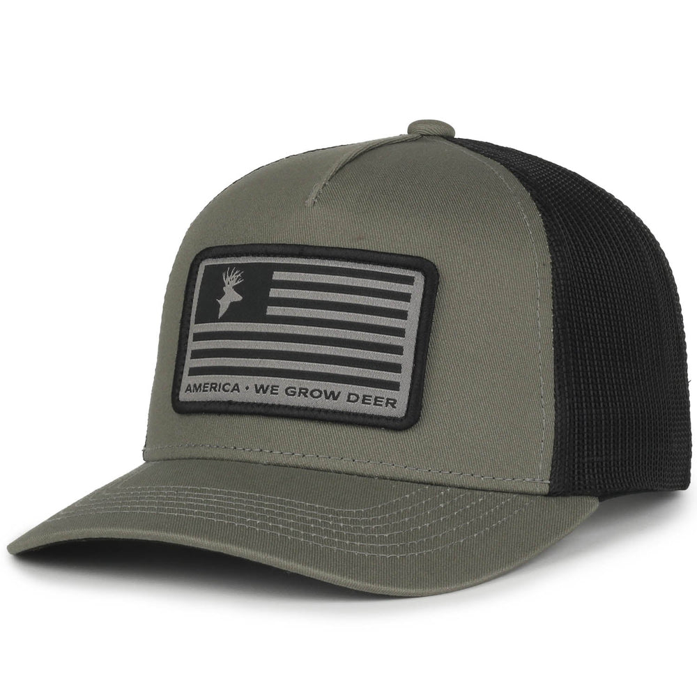 The Rural Cloth "We Grow Deer Flag Hat" in olive and black features an American flag design with a deer logo on the front patch. The text under the flag reads, "America - We Grow Deer, Big Bucks." It has a mesh back and an adjustable snap closure.