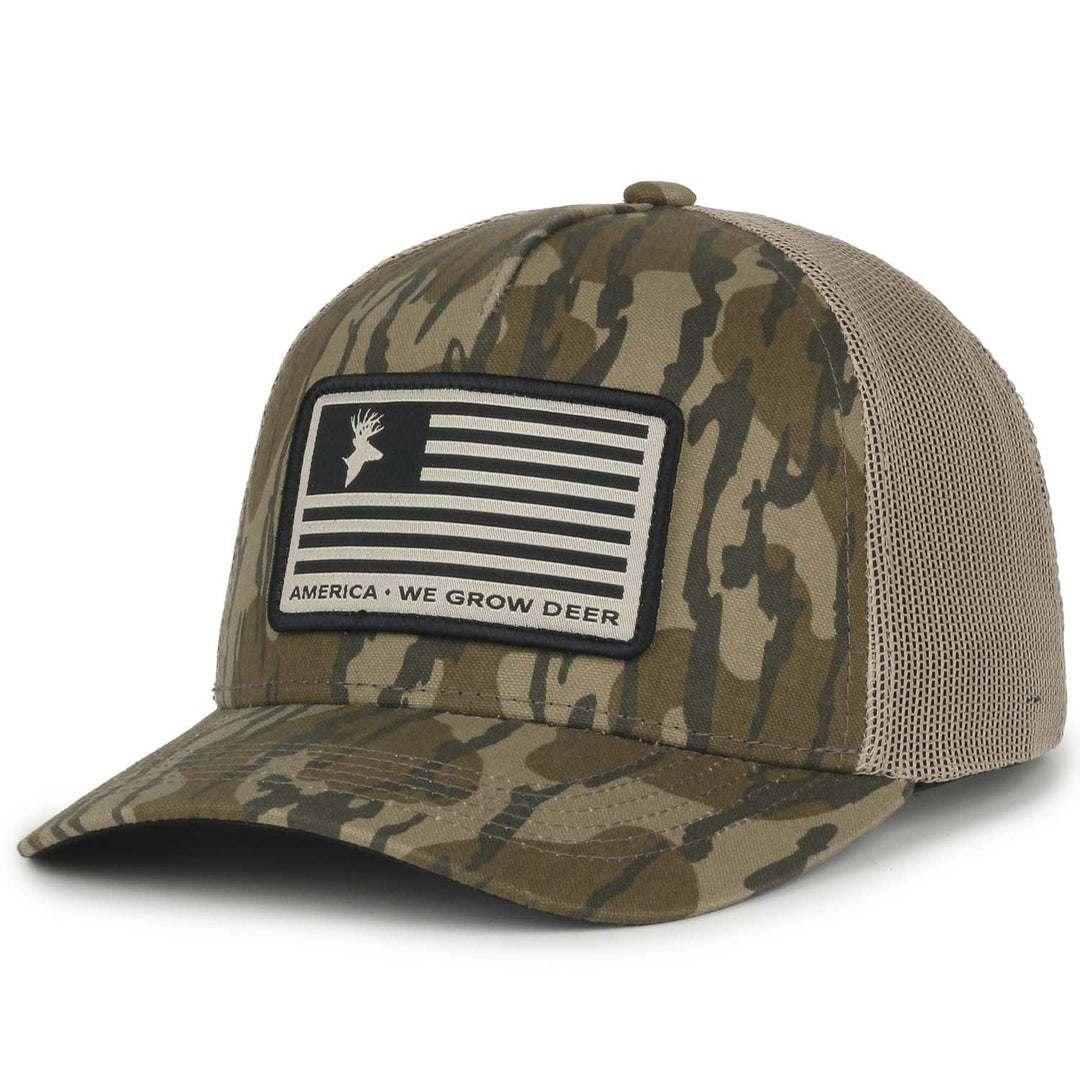 The We Grow Deer Flag Hat by Rural Cloth features a camouflage pattern in Bottomland. It has a mesh back, and the front showcases a patch with an American flag where the stars are replaced by a stylized deer head. Below the flag, the text reads "AMERICA - WE GROW DEER and big bucks.