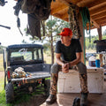 A man wearing the We Grow Deer snapback hat from Rural Cloth, camo pants, and a dark t-shirt sits on a white cooler on a wooden deck. Behind him, an off-road vehicle and various hunting gear hint at his passion for American deer hunting. The deck is attached to a wooden structure with hooks and shelves.