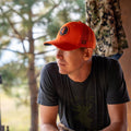 A man wearing the We Grow Deer Hat by Rural Cloth, featuring an orange snapback with an emblem, and dressed in a dark t-shirt, looks to the side. The background displays out-of-focus greenery and a hanging camouflage jacket, encapsulating the spirit of an American deer hunting outdoorsman.