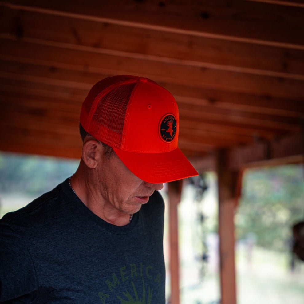 A man sporting the We Grow Deer Hat by Rural Cloth, distinguished by its bright red hue and black circular emblem, glances downward. He stands beneath a rustic wooden structure with a blurred background, embodying the spirit of an outdoorsman. His dark t-shirt with partially visible text adds to the authentic essence of American deer hunting culture.