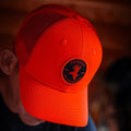 An outdoorsman is seen wearing the We Grow Deer Hat by Rural Cloth, an orange snapback cap with a mesh back. The hat features a circular patch on the front displaying a deer head silhouette alongside the words America We Grow Deer. The person's face remains hidden from view.