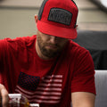 A bearded man wearing a Rural Cloth's We Grow Whiskey - 7-Panel hat, which features a red and black design with the 