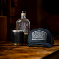 The image shows a black YETI tumbler, a bottle of whiskey, and the We Grow Whiskey Hat-Blackout by Rural Cloth, featuring an adjustable snapback closure and adorned with a white patch that reads 