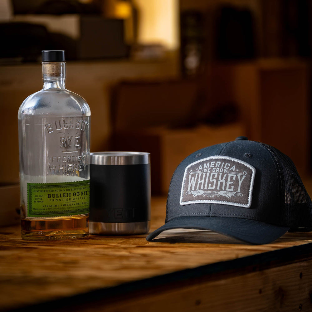 A bottle of Bulleit Rye whiskey, a Yeti tumbler, and a black We Grow Whiskey Hat-Blackout by Rural Cloth featuring an "Adjustable Snapback Closure" and "Whiskey" patch sit on a wooden surface. The background is softly blurred and suggests an indoor setting with warm lighting.