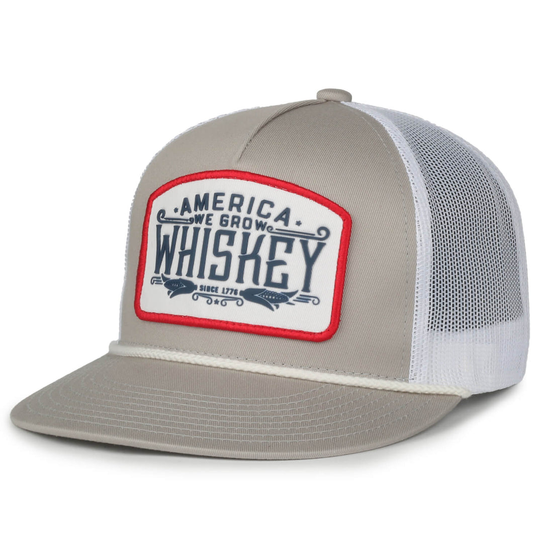 The We Grow Whiskey Hat - Flat by Rural Cloth is a stylish baseball cap featuring beige twill front panels and a white mesh back. The front panel is adorned with a stitched patch that boldly displays the text "America We Grow Whiskey," accented with a red border. The matching beige brim and convenient mesh back snapback closure complete the look.