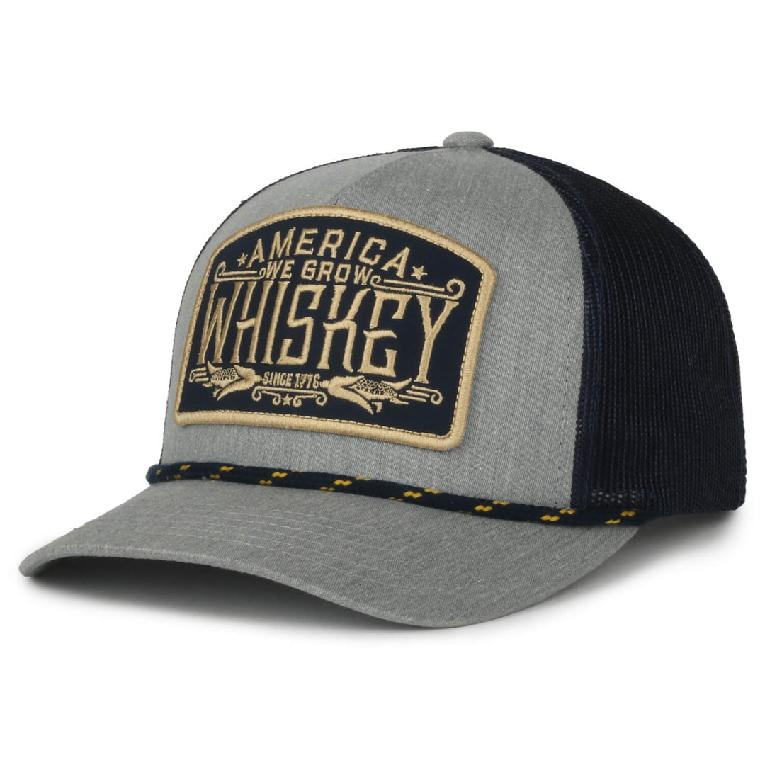 Introducing the We Grow Whiskey Hat-Gray by Rural Cloth: a stylish blue and black trucker hat with a mesh back and an adjustable snapback closure. The front features a distinctive patch displaying "America, We Grow Whiskey" in white and tan letters. This unique whiskey hat is adorned with braided detail near the bill and decorative stitching on the visor for added flair.