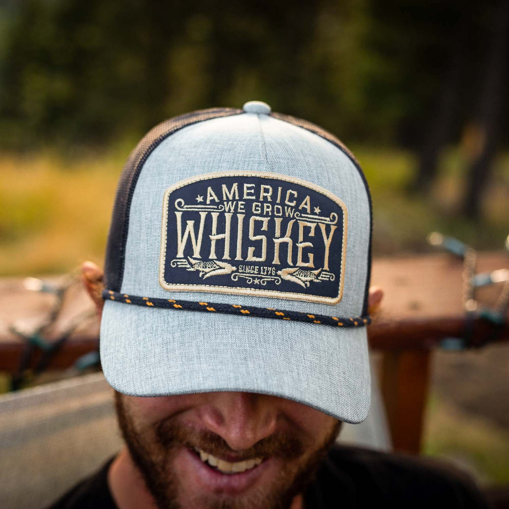 A close-up of a bearded person smiling while wearing the "We Grow Whiskey Hat-Gray" by Rural Cloth, which features a patch that reads "America: We Grow Whiskey Since 1776" and an adjustable snapback closure. The background is blurred, highlighting outdoor vegetation and what appears to be wooden furniture.