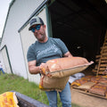 A man wearing a green We Hunt Deer Tee by Rural Cloth, along with a baseball cap and sunglasses, is carrying bags of animal feed near a barn. He is placing one bag into a truck bed, with additional bags stacked nearby. The scene takes place outdoors on a grassy area.