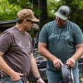 Two men are standing outside near some machinery, both wearing baseball caps and the 