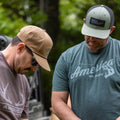 Two men wearing caps and Rural Cloth's We Hunt Deer Tee, made from super soft cotton, stand outdoors with focused expressions. One man is looking down while the other gazes off to the side. Trees are visible in the background, suggesting they are in a forested area, likely showcasing high-quality hunting apparel made in the USA.