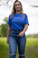 A woman with long blonde hair is walking outdoors on a grassy path, wearing a comfortable and durable Rural Cloth Western Tee and distressed jeans. The blue T-shirt features white text that reads 