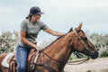 A person wearing a comfortable and durable gray Rural Cloth Western Tee and a baseball cap is sitting on a brown horse. The individual, donning the Western Tee, looks off to the side with one hand on the horse's neck. In the background, trees and an open field evoke scenes reminiscent of the American cattle industry.