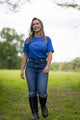 A woman is walking on a grassy field in a Rural Cloth Western Tee, blue jeans, and black boots. Trees and greenery are visible in the background under an overcast sky. She smiles while looking ahead, conveying the spirit of the American cattle industry with her comfortable and durable attire.