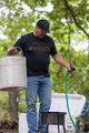 A man wearing a Whiskey Tee from Rural Cloth, blue jeans, and a black cap holds a basket in his left hand and a hose in his right, standing next to an open white cooler. The background shows green trees and a natural outdoor setting.