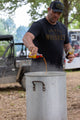 A whiskey aficionado wearing the Whiskey Tee by Rural Cloth and a cap is pouring a liquid from two bottles into a large, steaming metal container outdoors. In the background, a vehicle rests on the dirt surface, surrounded by trees and greenery.