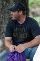 A whiskey aficionado in a Whiskey Tee by Rural Cloth and a black baseball cap is sitting outdoors. He holds a purple mesh bag and gazes to his left, surrounded by trees and greenery in the background.