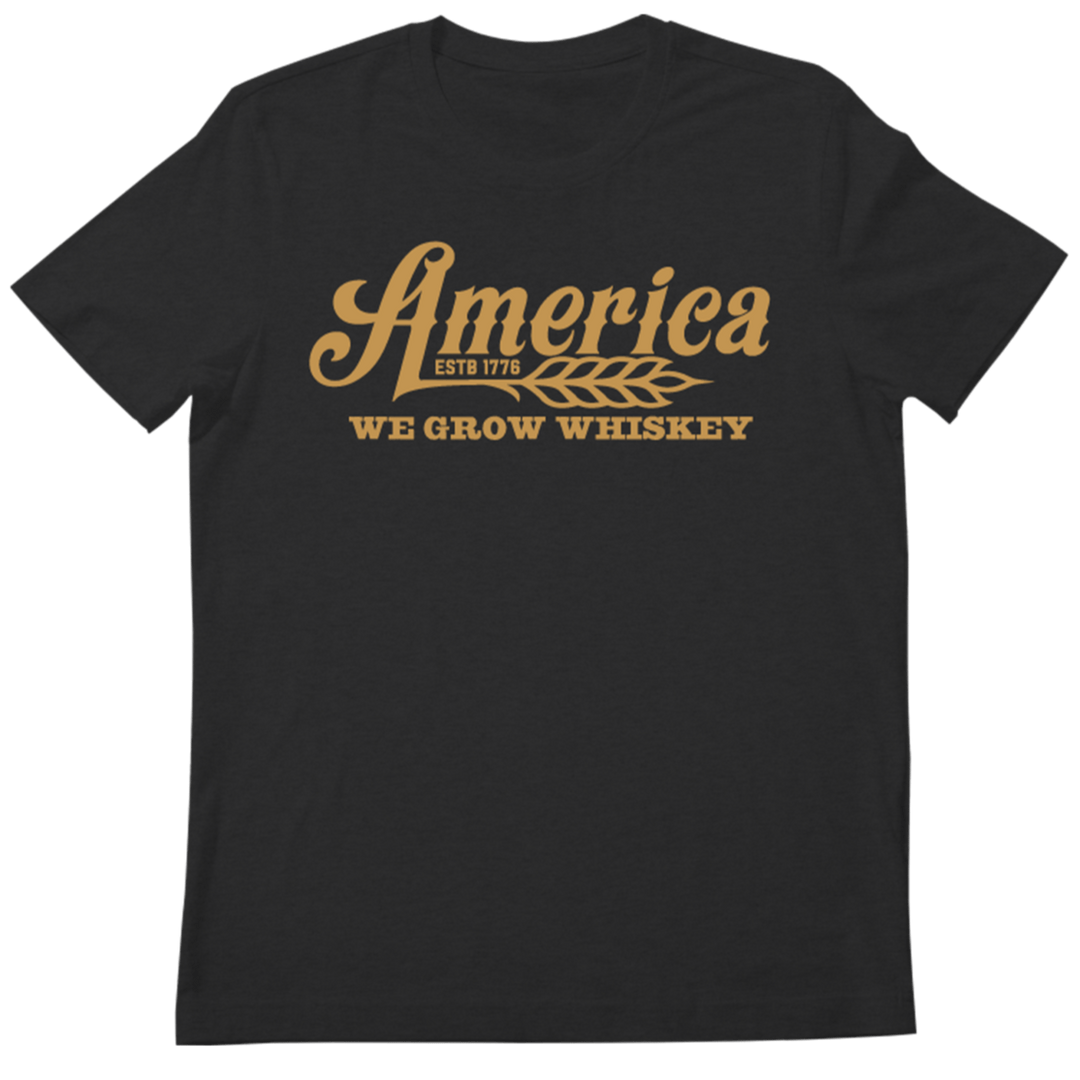 The 76 Wheat Tee-Black by Rural Cloth features orange text that reads "America" above a graphic of a wheat stalk, with "ESTD 1776" in small letters within the stalk, and "WE GROW WHISKEY" below the graphic. This black t-shirt celebrates America's rich heritage of whiskey production.