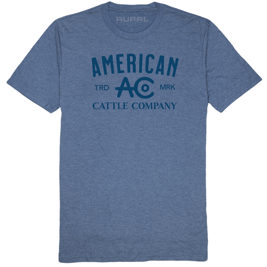 A blue ACC Brand Tee featuring "American Cattle Company" in dark blue letters, with the abbreviations "TRD" and "MRK" flanking the central "AC" logo. Made from super soft combed ring-spun cotton, the brand name "Rural Cloth" is visible on the inside of the collar.