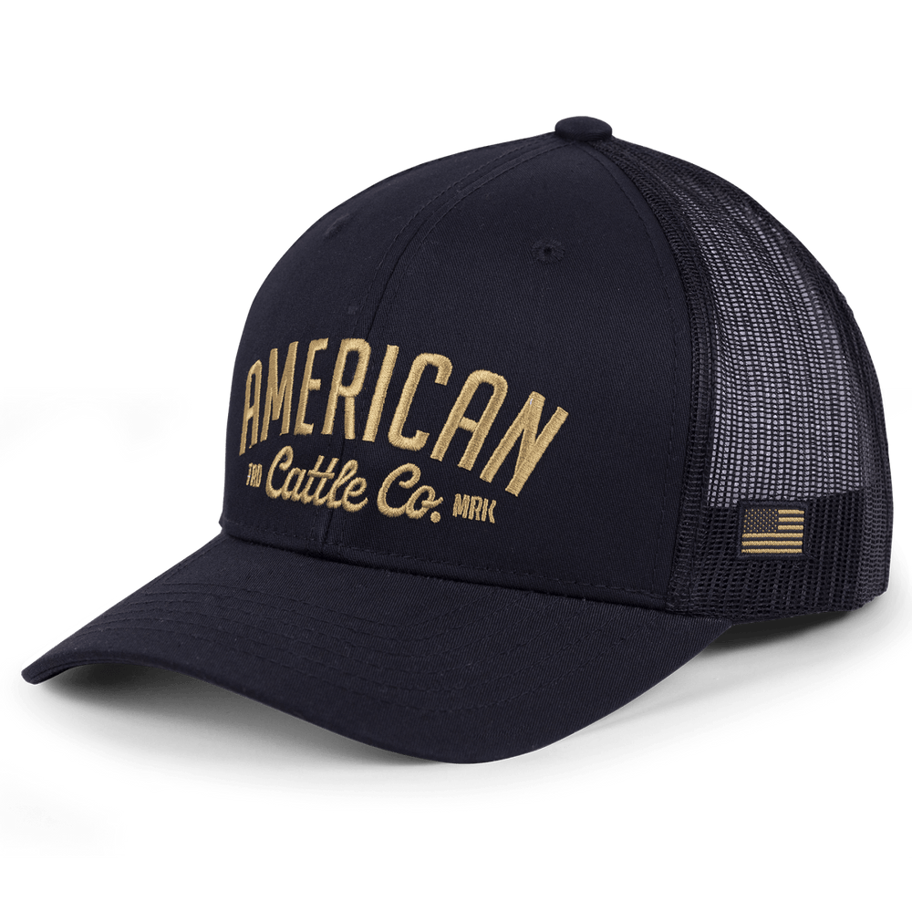 The ACC Embroidered Hat by Rural Cloth is a black baseball cap with "American Cattle Co." embroidered on the front in tan lettering. The back half features black mesh and an adjustable snapback closure, along with a small American flag patch on the side.
