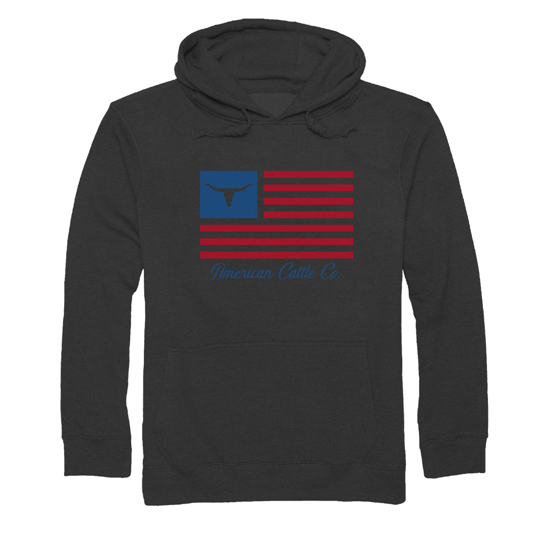 The ACC Flag Pullover-Charcoal Heather by Rural Cloth is a dark gray hooded sweatshirt featuring a graphic design of a stylized American flag on the front. The flag showcases a blue square with a bull skull silhouette and red stripes, with the text "American Cattle Co." in blue below, celebrating American cattle ranching.
