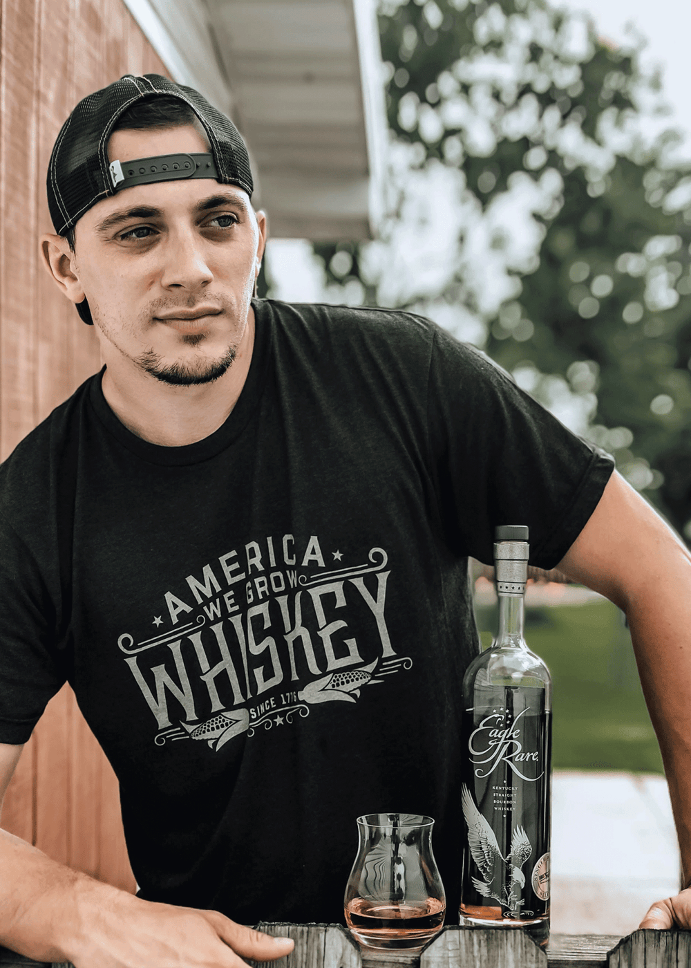 A man wearing a Rural Cloth "America We Grow Whiskey Tee" with "America Powered By Whiskey" printed on it is standing outdoors by a wooden surface. Next to him on the surface are a bottle of whiskey and a glass partially filled with whiskey.