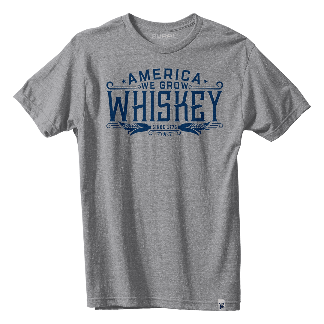 The "America We Grow Whiskey Tee - Gray" from Rural Cloth is a premium short-sleeved shirt featuring a blue graphic design that reads "America We Grow Whiskey Since 1776" with decorative elements around the text, proudly made in the USA.