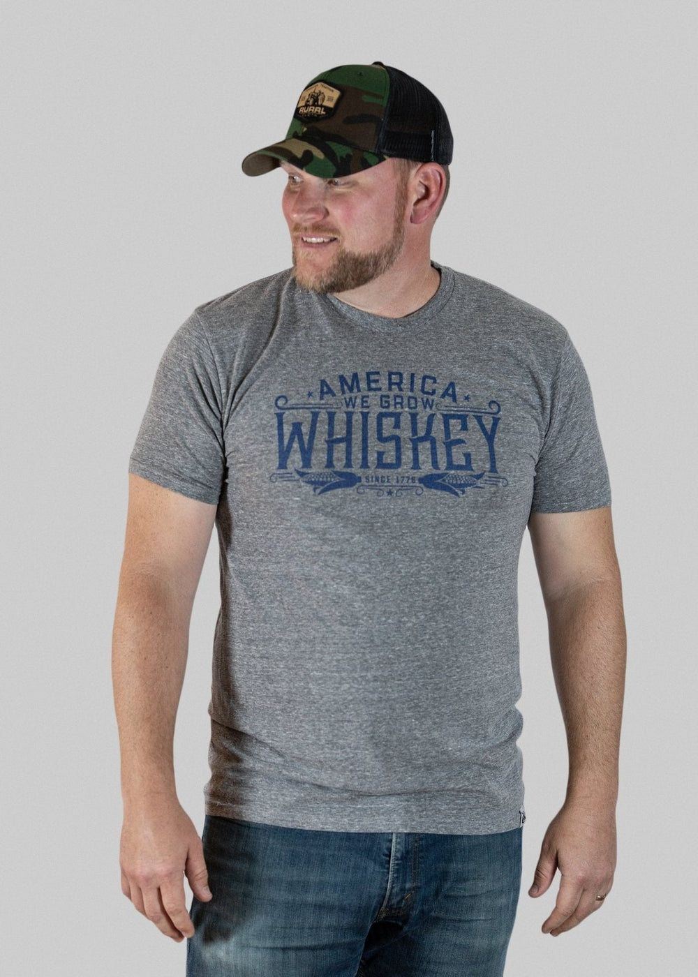 A man wearing a Rural Cloth "America We Grow Whiskey" Tee in gray and a camo baseball cap stands against a light gray background. The whiskey shirt pairs well with his faded blue jeans, reflecting that classic, made-in-USA style. He has a trimmed beard and looks to his right.