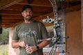 A man wearing an America We Hunt Elk Tee in Military Green Frost from Rural Cloth and a cap stands under a wooden structure, holding a beverage can in one hand. A compound bow is visible in the foreground. The setting appears to be outdoors, with part of a building and some greenery in the background.