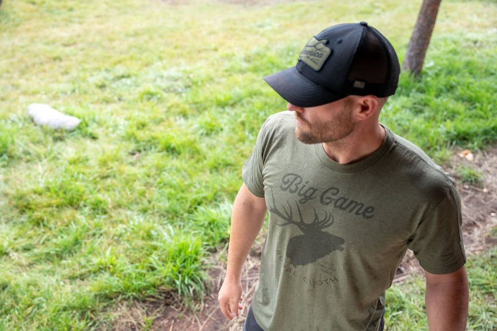 A bearded man stands on grass, looking to the side while wearing a Military Green Frost Big Game Tee from Rural Cloth featuring a deer graphic and paired with a black cap. He is posed outdoors near patches of greenery and dirt, seemingly ready for the next elk bugling season.