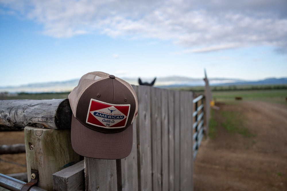 A brown and tan Billboard Hat by Rural Cloth, adorned with the "American Cattle Co." logo, rests on a wooden fence post. Featuring an adjustable snapback closure and a breathable mesh back, the cap sits against a backdrop of open fields and distant mountains under a partly cloudy sky. A horse's ears are visible behind the fence.