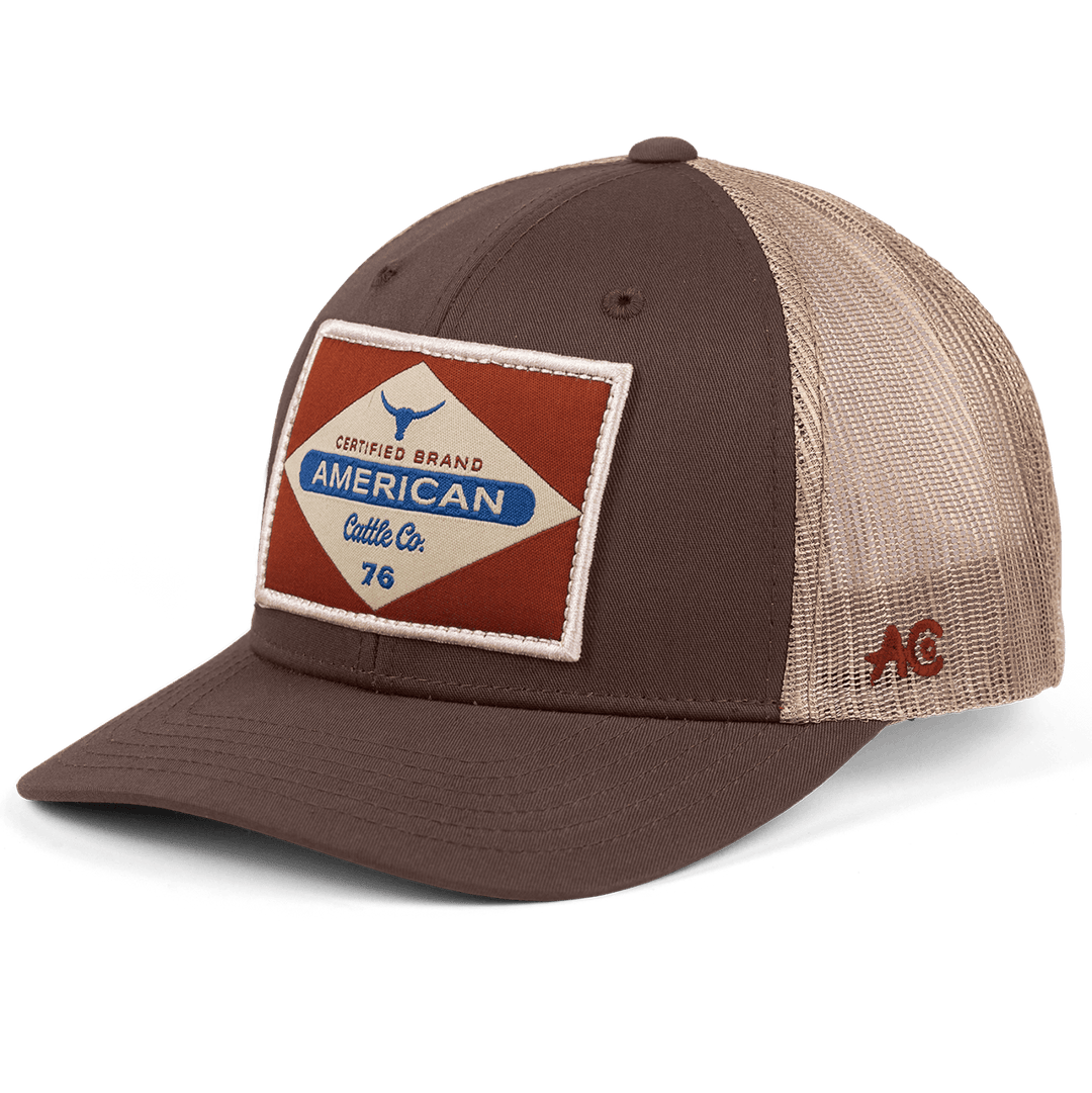 The "Billboard Hat" by Rural Cloth is a brown and beige trucker hat with a breathable mesh back. The front panel features a diamond-shaped patch with the text "Certified Brand American Cattle Co. 76" along with a small bull illustration. The side is stitched with the initials "AC" in red, and it includes an adjustable snapback closure for a perfect fit.