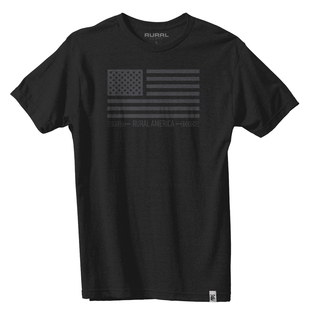 The Black Out Flag Tee by Rural Cloth is a comfortable black T-shirt adorned with a dark gray American flag design and the text "RURAL AMERICA" at the center. This shirt features the brand name "Rural" on the inside collar and a small white logo tag attached to the bottom hem, showcasing American pride.