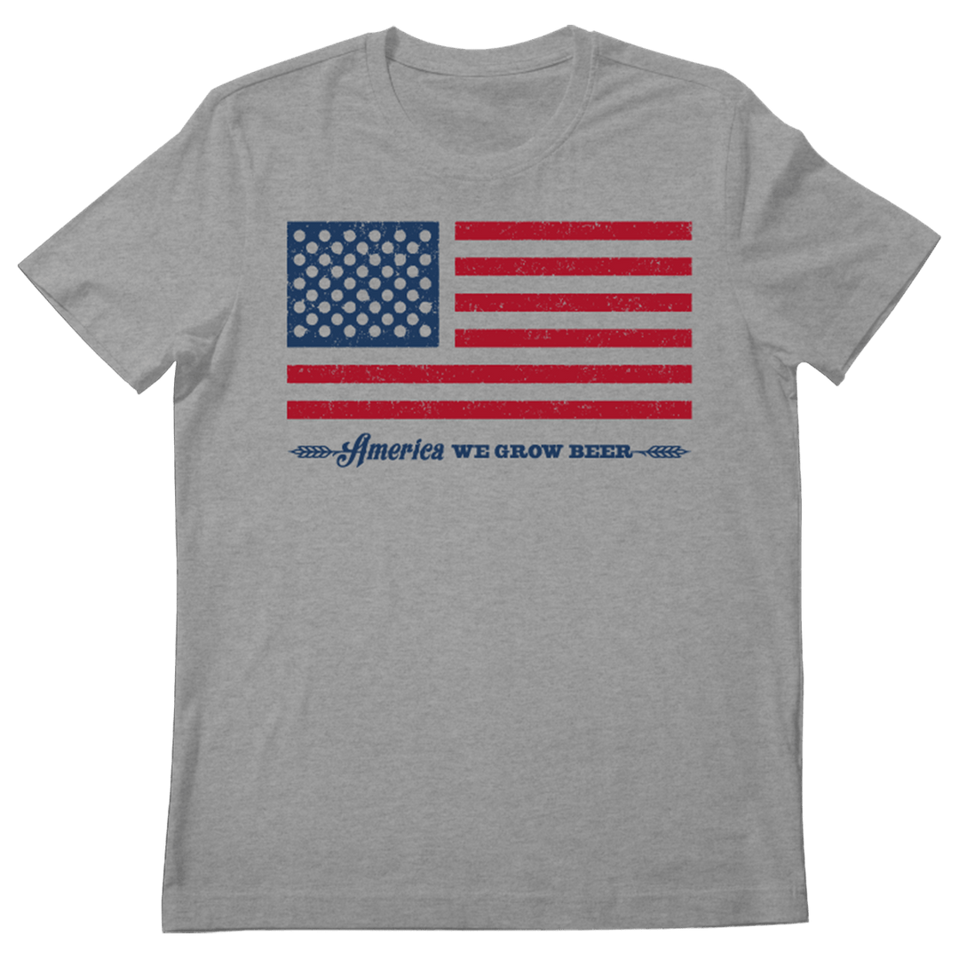 The Bottle Cap Tee by Rural Cloth is a gray T-shirt featuring a stylized American flag with blue stars and red stripes. Beneath the flag, the text reads, “America We Grow Beer,” accompanied by two small wheat designs. This tee is ideal for patriotic celebrations and showcases American beer culture.