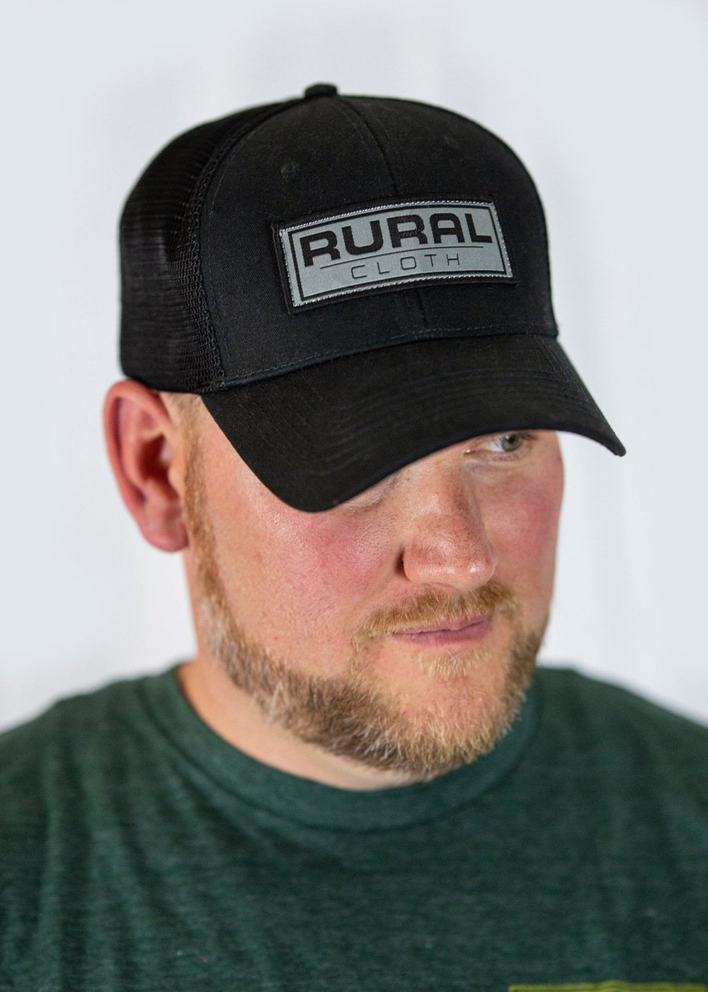 The image shows a bearded man wearing a dark green t-shirt and the Brandmark Hat from Rural Cloth, an adjustable snapback black baseball cap featuring a rectangular patch on the front that reads "RURAL CLOTH." He is gazing slightly to the side against a plain, light-colored background.