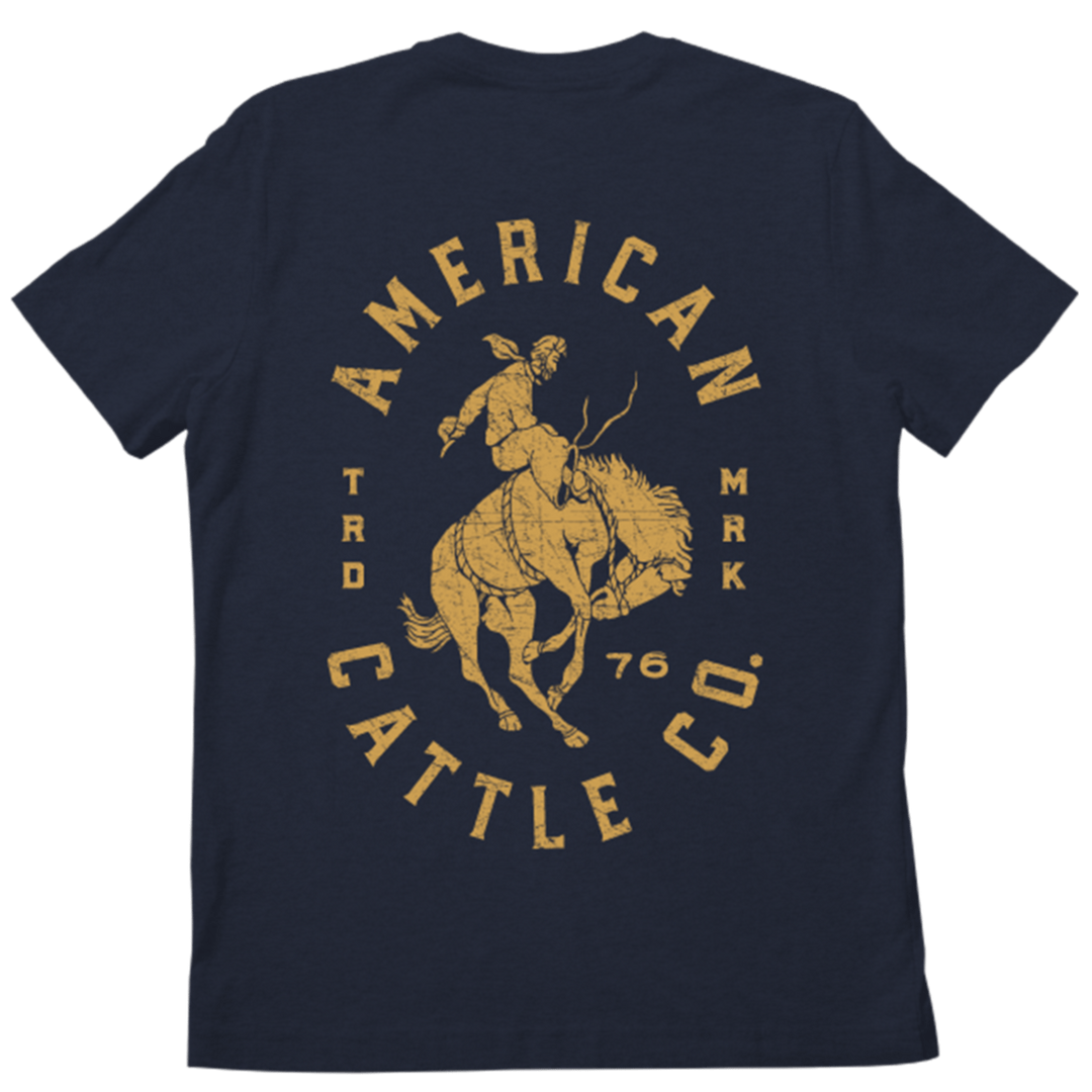 The image shows the back view of a navy blue Bronco TM Tee by Rural Cloth. Printed on it in mustard yellow is a graphic of a cowboy riding a bucking horse, with the text "AMERICAN CATTLE CO." and "TRD MRK 76" arranged around the graphic, evoking classic American ranching vibes.