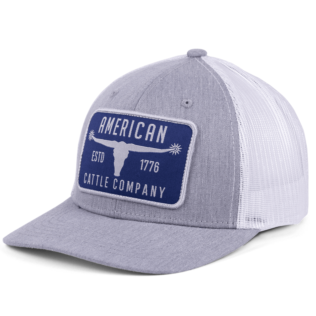 The "Bull Spurs Hat-Gray with Blue Patch" by Rural Cloth is a stylish gray and white trucker hat featuring a mesh back and an adjustable snapback closure. Adorning the front is a blue patch with white text and an image of a longhorn cow skull, bearing the words: "American Cattle Company," "ESTD 1776." This hat is perfect for fans of cattle and country living.
