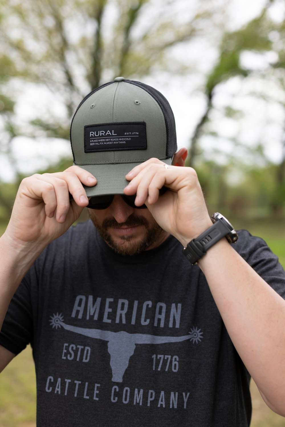 A person wearing a gray cap and a black "Rural Cloth Bull Spurs Tee-Black Frost" shirt adjusts the brim of the cap with both hands. The premium weight fabric of the shirt hints at its quality, and the background features blurred greenery.