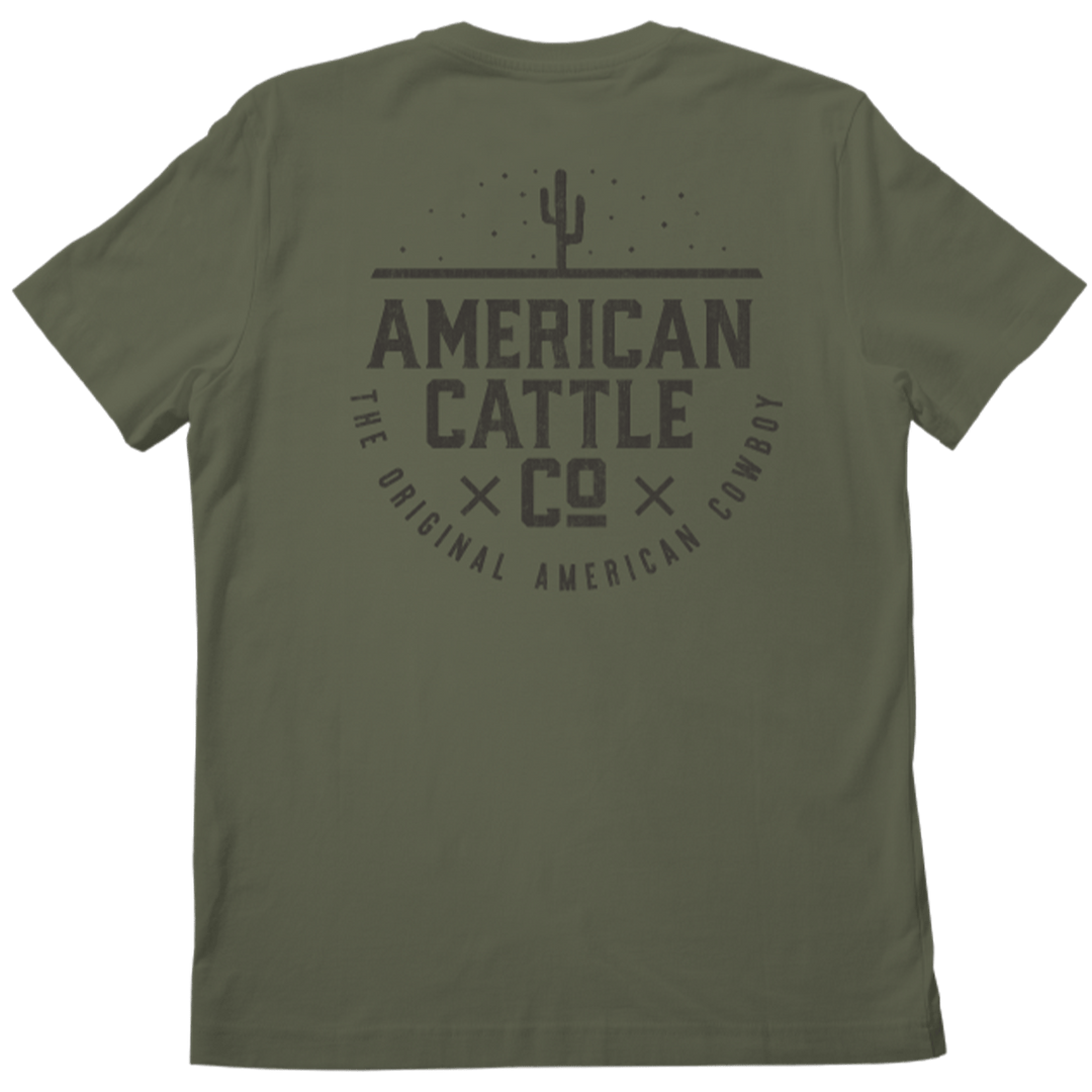 This image showcases the back of an olive green Cactus Tee from Rural Cloth, featuring a striking graphic design. The artwork includes a cactus illustration and bold black text that reads "American Cattle Co" and "The Original American Cowboy," evoking the rugged essence of a desert landscape.