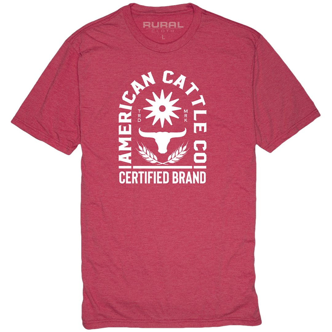 The Certified Tee by Rural Cloth is a red T-shirt featuring a white graphic design. The design includes the text "American Cattle Co" surrounding a sun with rays and a longhorn skull encircled by wheat stalks, celebrating ranching heritage. Below, it reads "Certified Brand." The shirt is available in size L.