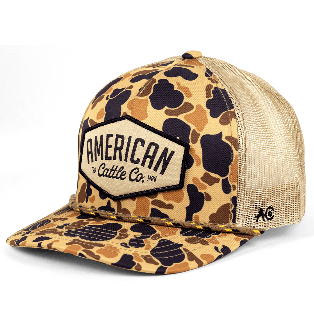 The Diamond Hat by Rural Cloth features a camouflage-patterned design with a beige mesh back and a front patch boldly displaying "American Cattle Co." To celebrate American heritage, the brim also sports matching camo, while a small logo is embroidered on the side panel.