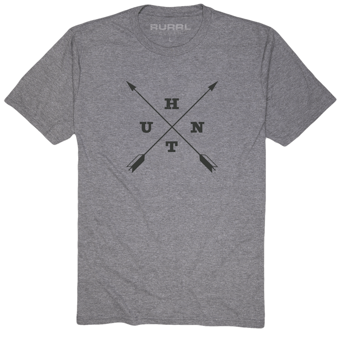 The Hunt Tee by Rural Cloth in heather gray showcases a unique design with two black arrows crossed in the center. The letters U, H, T, and N are strategically positioned between the arrows. Additionally, the word "Rural" is printed on the inner back collar, making it an ideal choice for hunters who crave adventure.